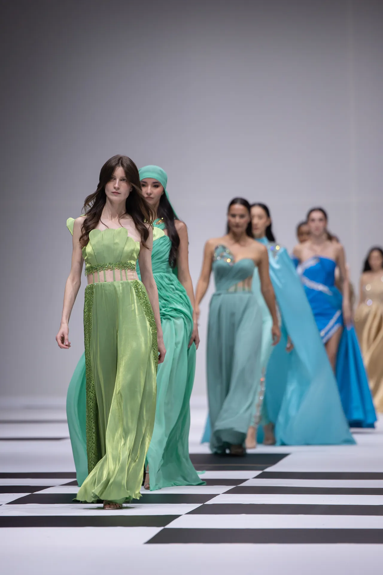 Front model walks the runway in a lime-green gown with sheer panels, followed by other models in pastel gowns on a black-and-white checkerboard runway.