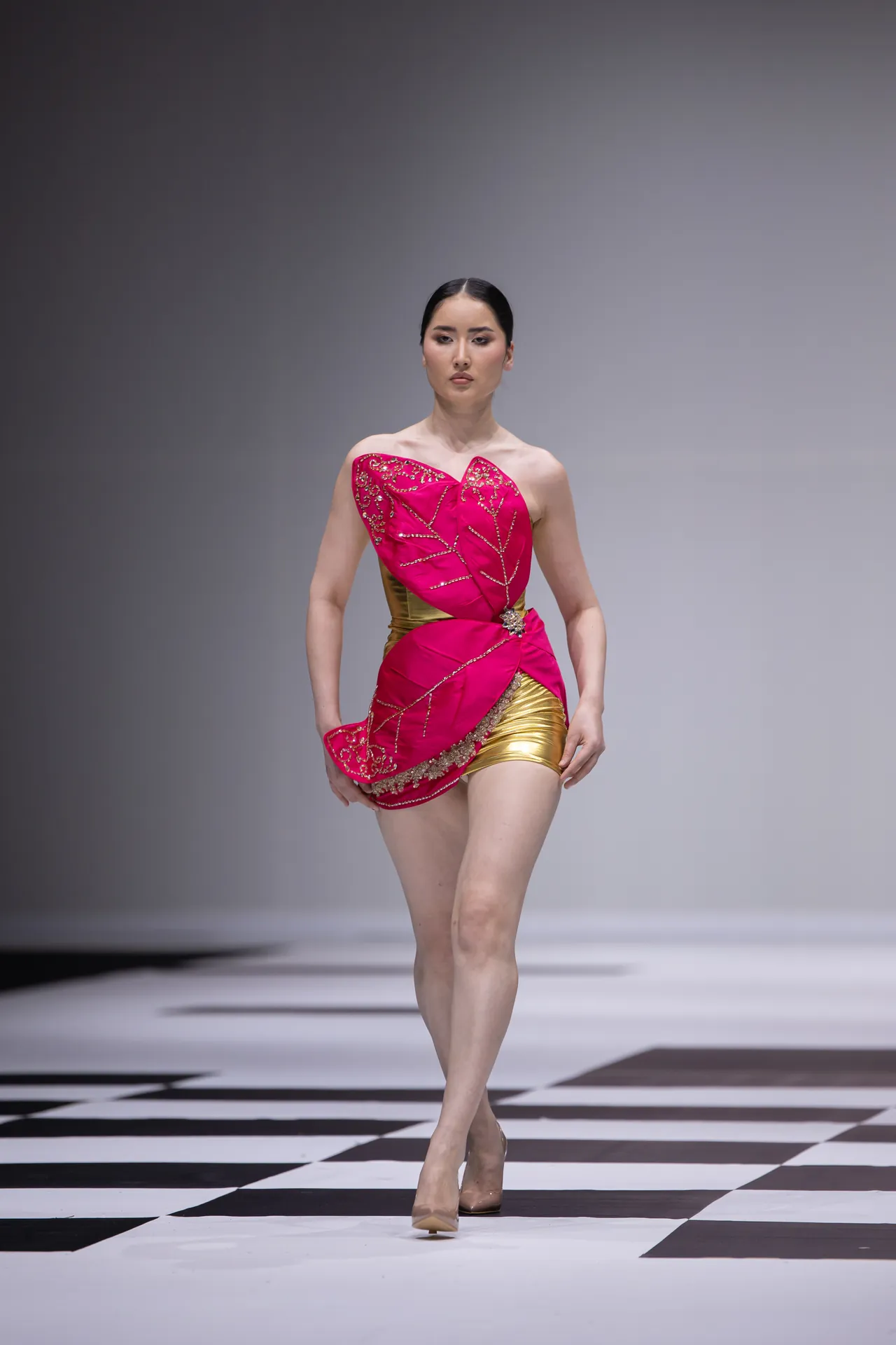Model walks the runway in a bright pink, strapless dress with gold embellishments and a gold waist, on a black-and-white checkered catwalk.