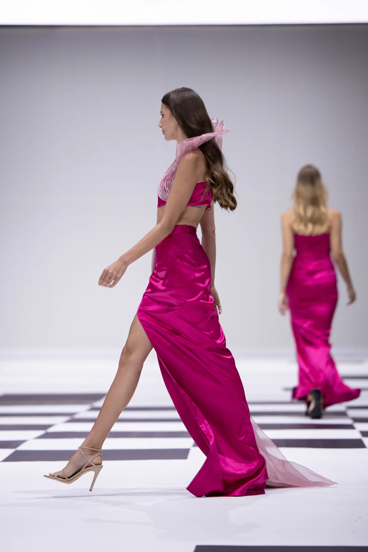 Model walking a fashion runway in a magenta satin two-piece outfit with a high slit, beige heels visible.