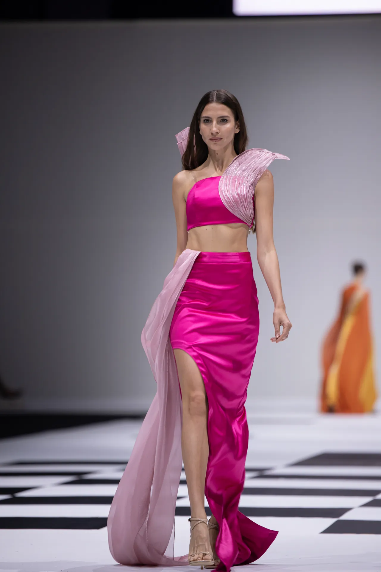 Model on a runway wearing a bright pink two-piece outfit with a sheer pink cape flowing at her side.
