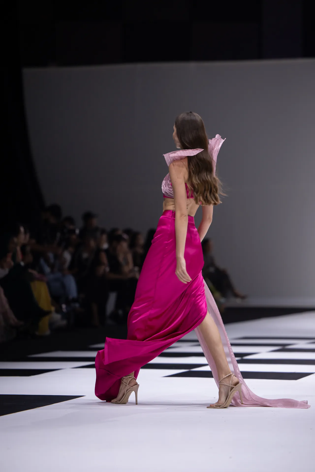 Model walking a fashion runway in a bright pink dress with ruffled pink shoulders and nude heels, audience blurred in the background.
