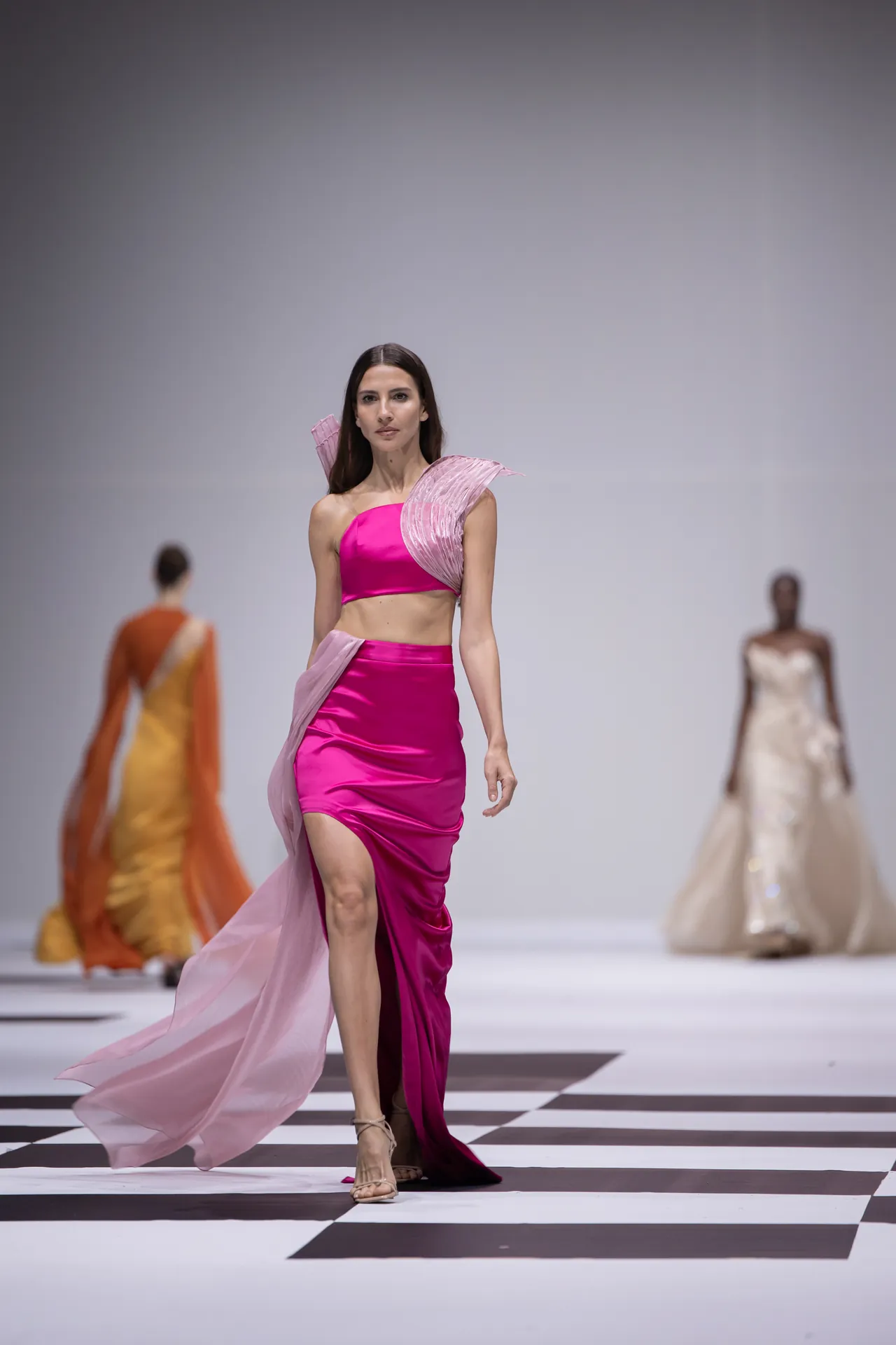 Female model walking a fashion runway in a hot-pink two-piece outfit with a pink flowing cape, on a black-and-white checkered floor.