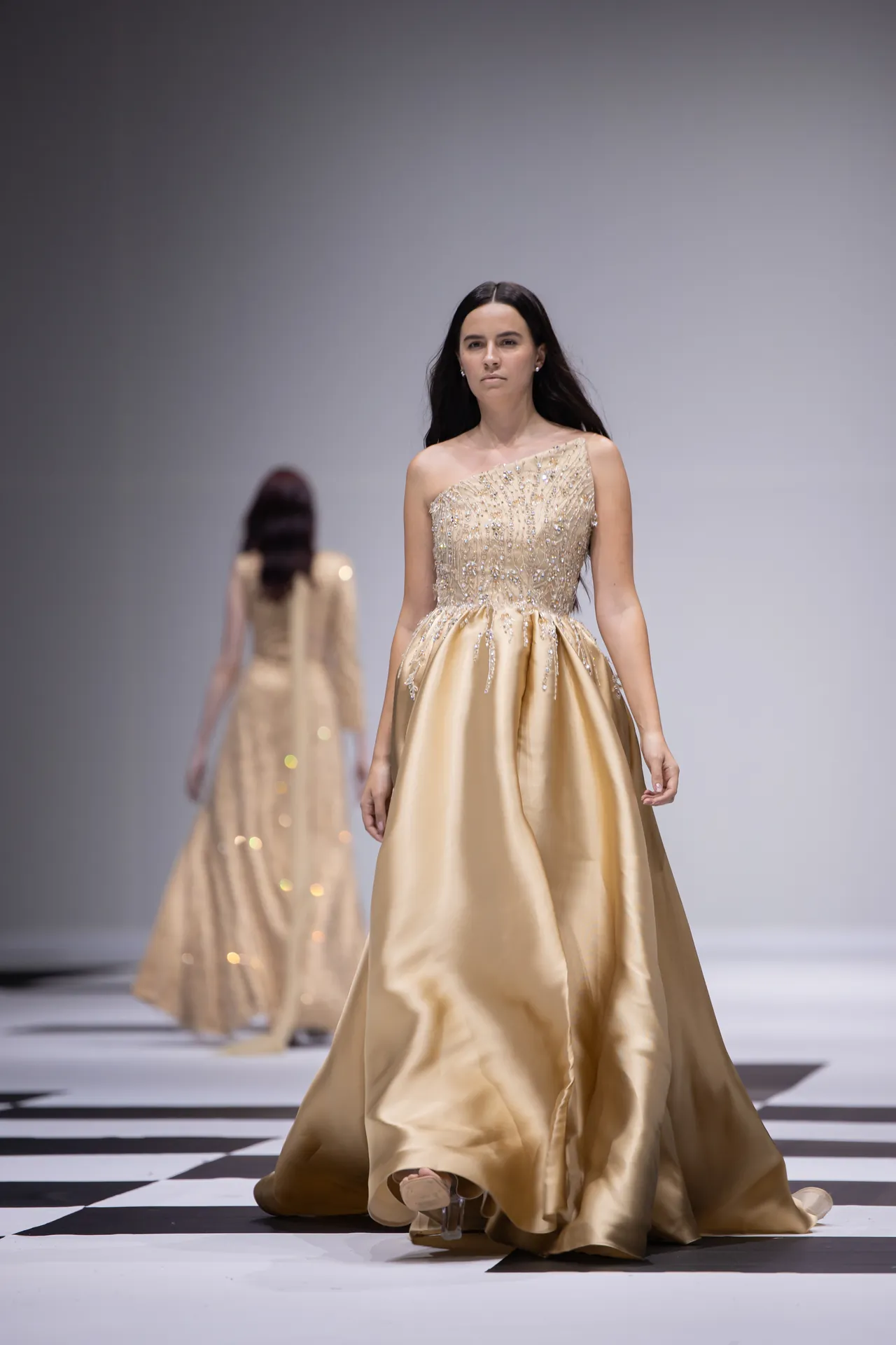 Model walks a gold embellished ball gown down a runway; other models in similar dresses blur in the background on a checkered runway.