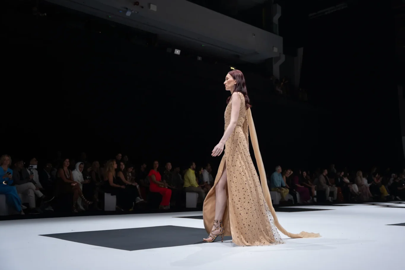 Model in a champagne-colored, embellished gown with a flowing cape walks the runway as the audience watches from the darkened backdrop.