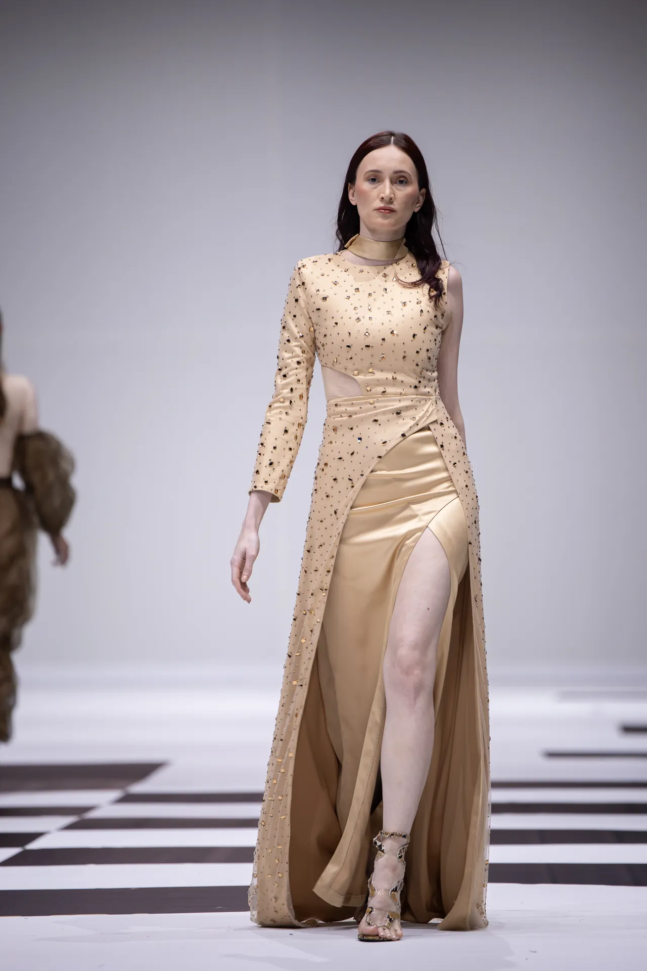 Model walks the runway in a champagne-colored gown with gold embellishments and a high front slit, beige cape-like overlay, and strappy heels.