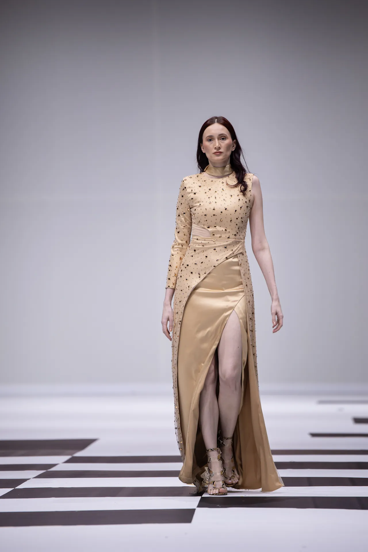 Model walking a fashion runway in a beige embellished gown with a thigh-high slit on a black-and-white checkerboard floor.