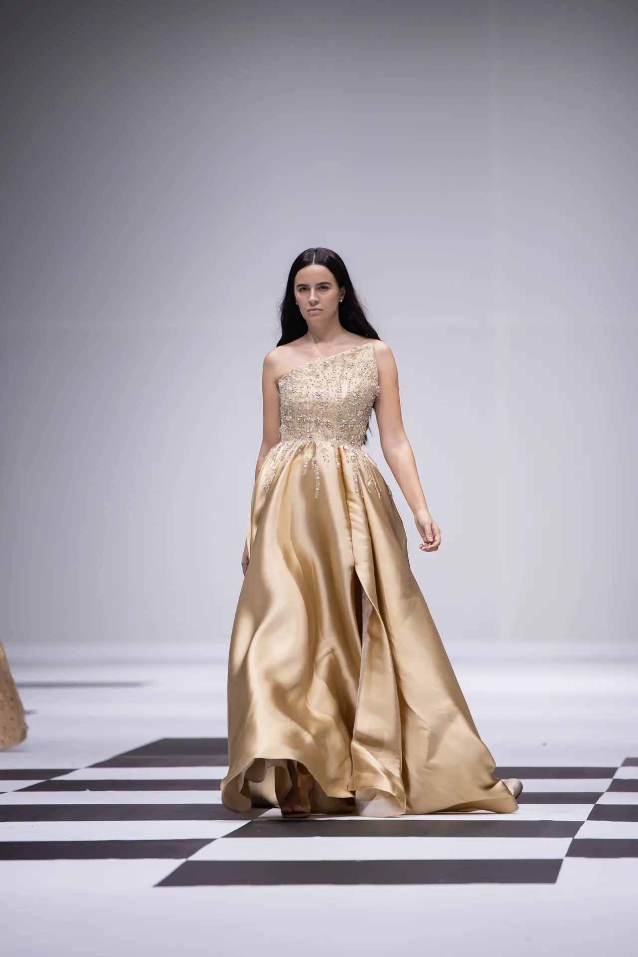 Model walking a runway in a gold, one-shoulder gown with a beaded bodice against a checkered floor and neutral backdrop.