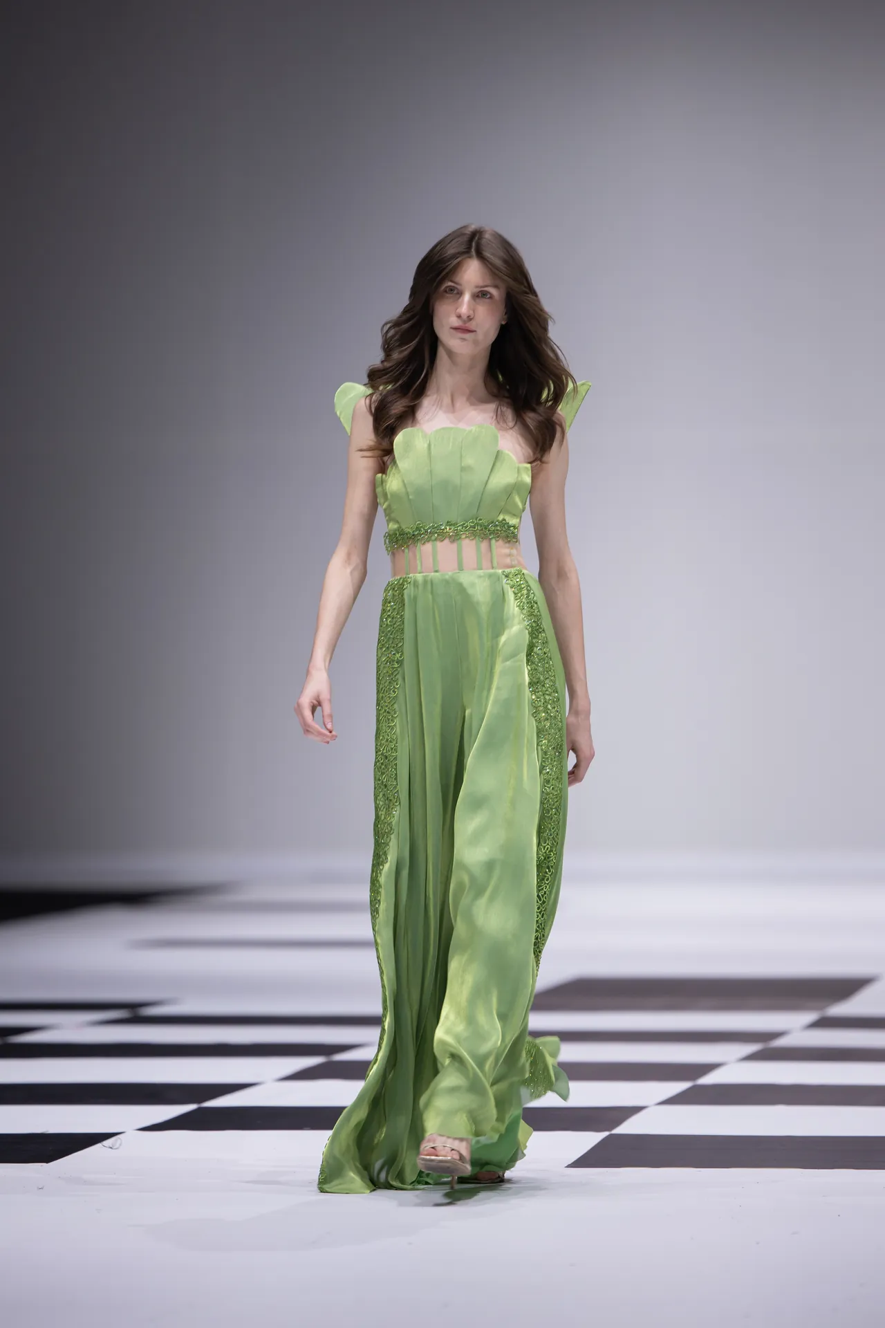 Female model walks the runway in a lime-green, flowing gown with sheer waist panels and sculpted bodice details.