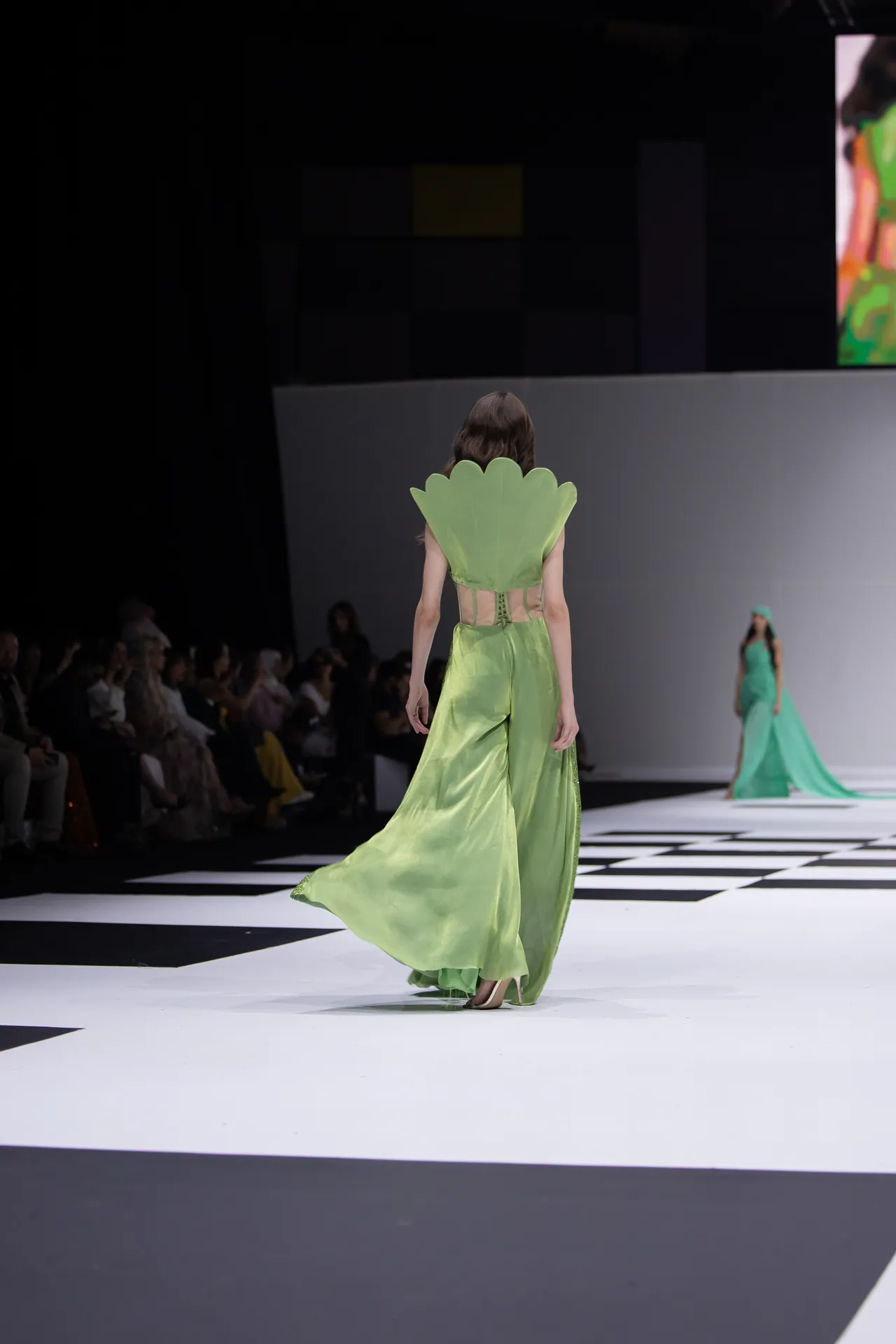 Model walks a bright green gown with a large scalloped leaf-like back on a fashion runway; audience seated along the sides.