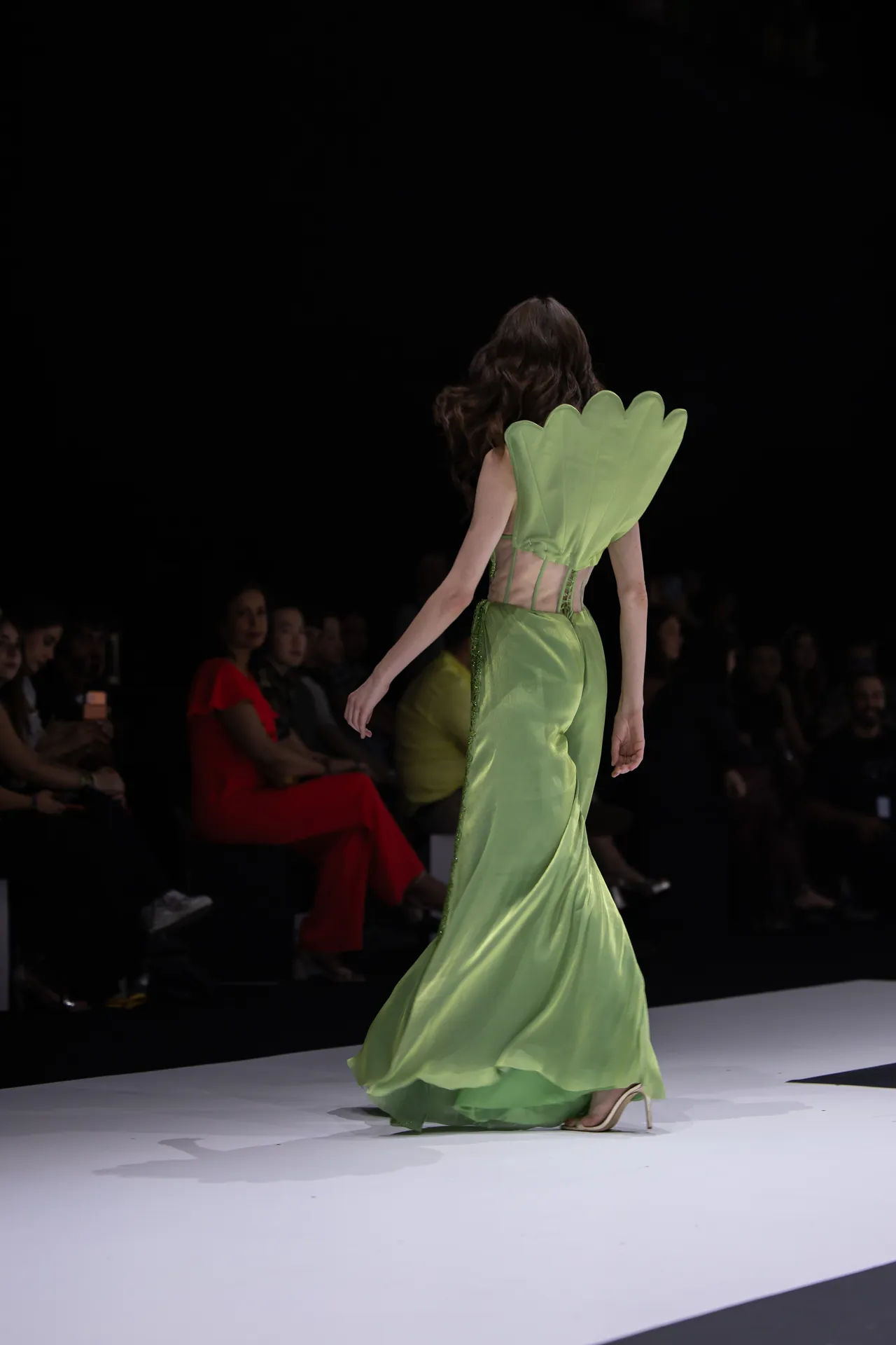 Runway model walking in a light green satin gown with large leaf-shaped back adornment, seen from behind on a white catwalk with a dark audience backdrop.