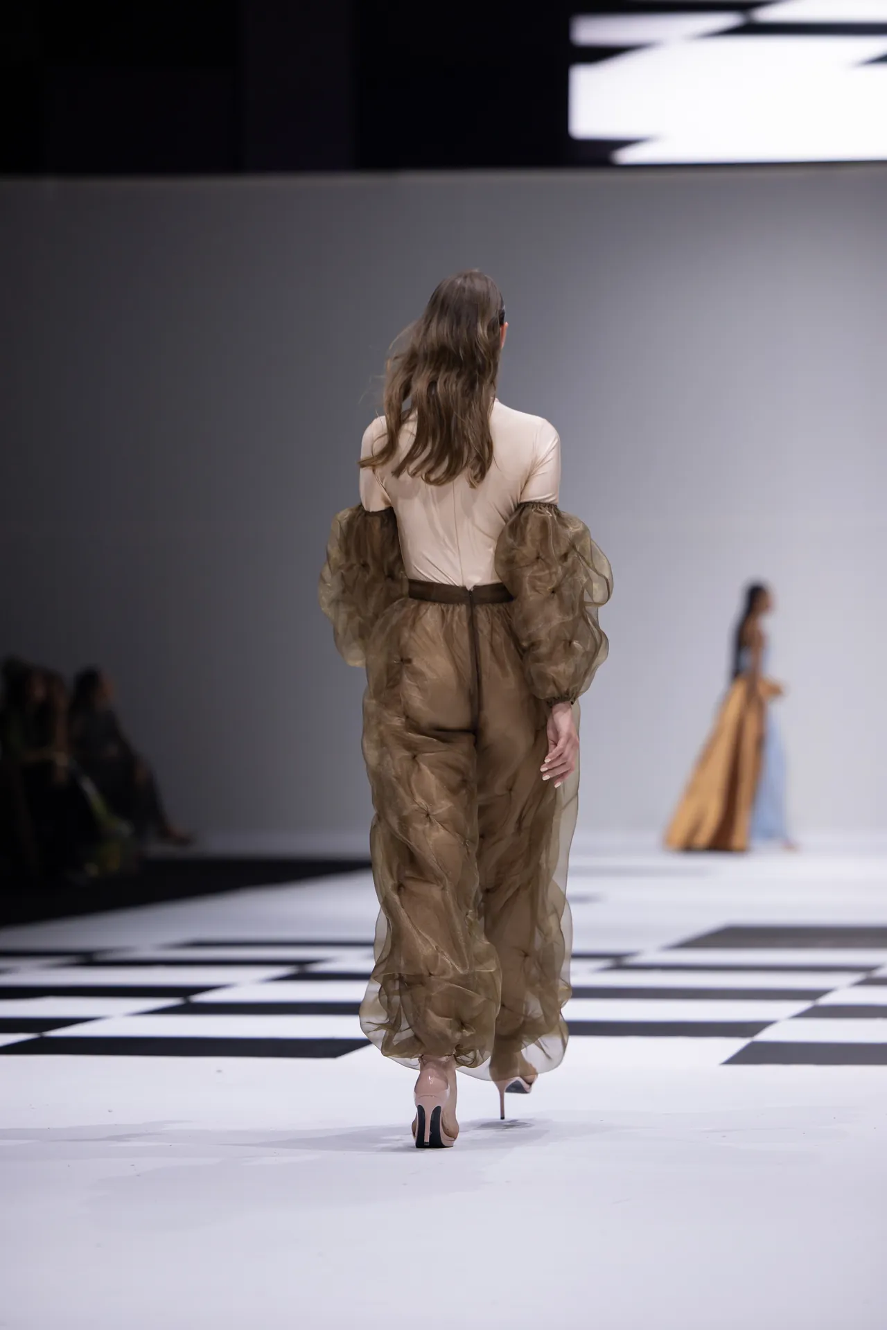 Back view of a fashion model walking the runway in a tan sheer jumpsuit with voluminous puff sleeves.