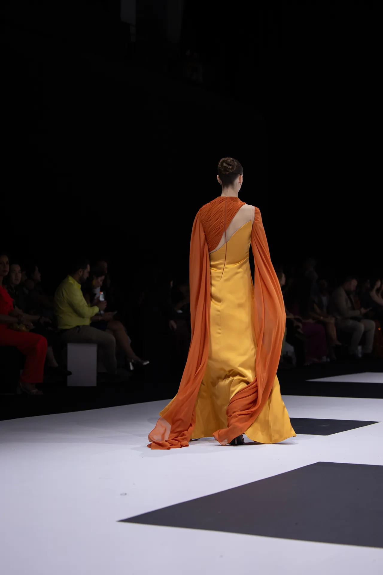 Back view of a model walking a fashion show in a yellow gown with an orange cape flowing behind her on a white runway distance to the dark audience.