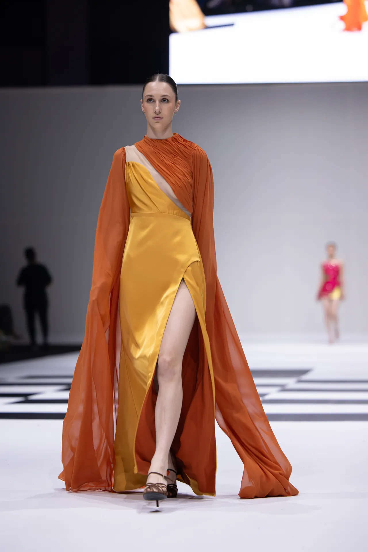 Model walking a runway in an orange and yellow color-block gown with a high slit and a sheer orange cape flowing behind her.