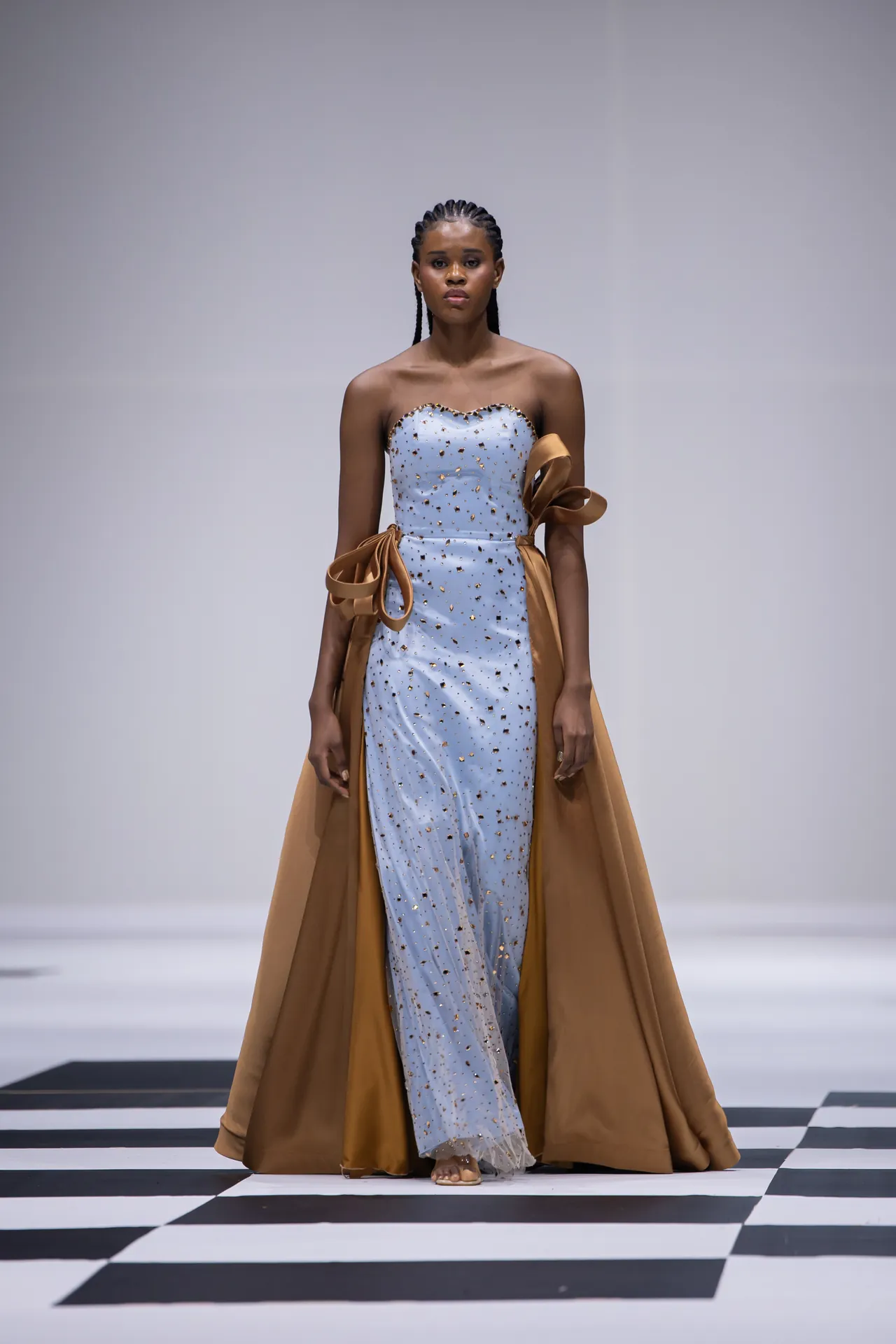 Model walks a fashion runway in a strapless light-blue gown studded with gold, paired with a brown cape.