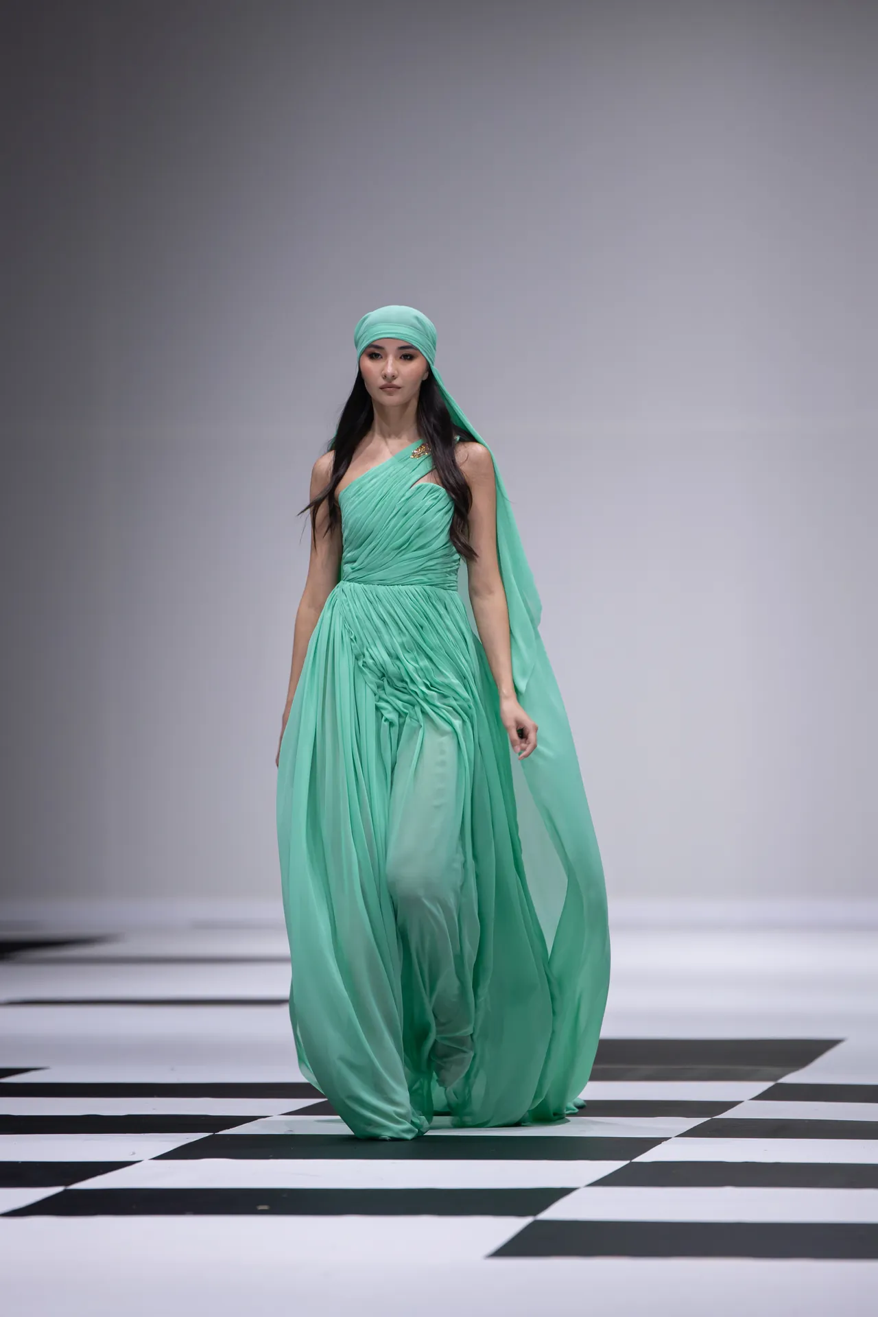 Model walking a fashion runway in a flowing mint-green gown with a matching head wrap and cape, on a black-and-white checkered floor.