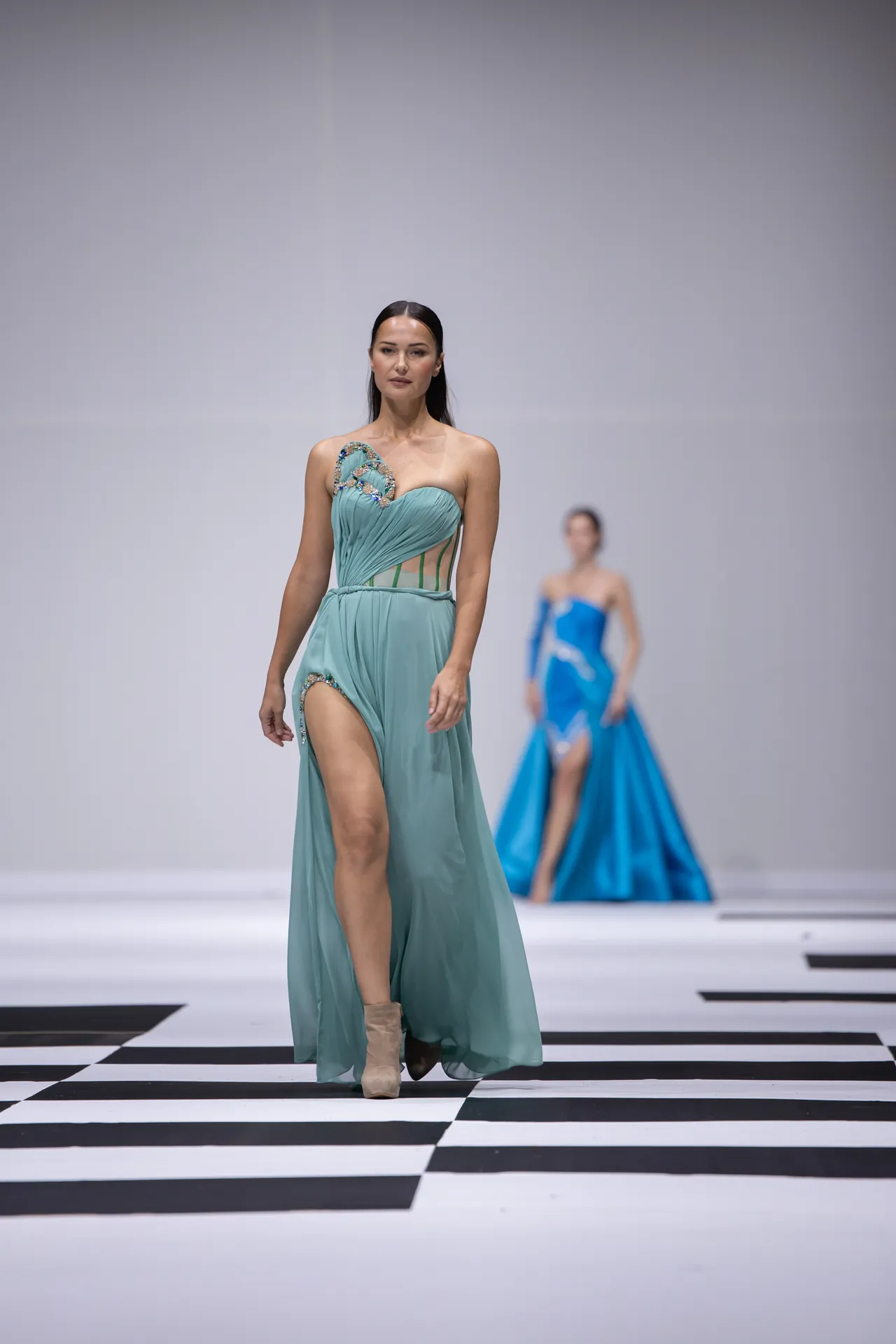 Runway model in a teal, one-shoulder gown with a high slit walking toward the camera; black-and-white checker floor.