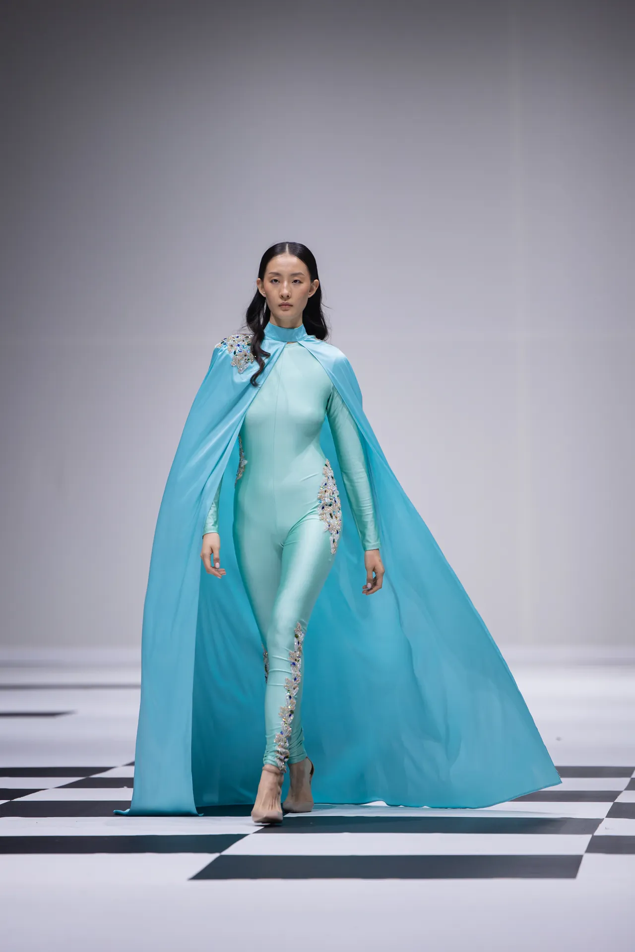 Model walks a runway in a turquoise cape and matching form-fitting bodysuit with jeweled embellishments on the shoulders and hips.