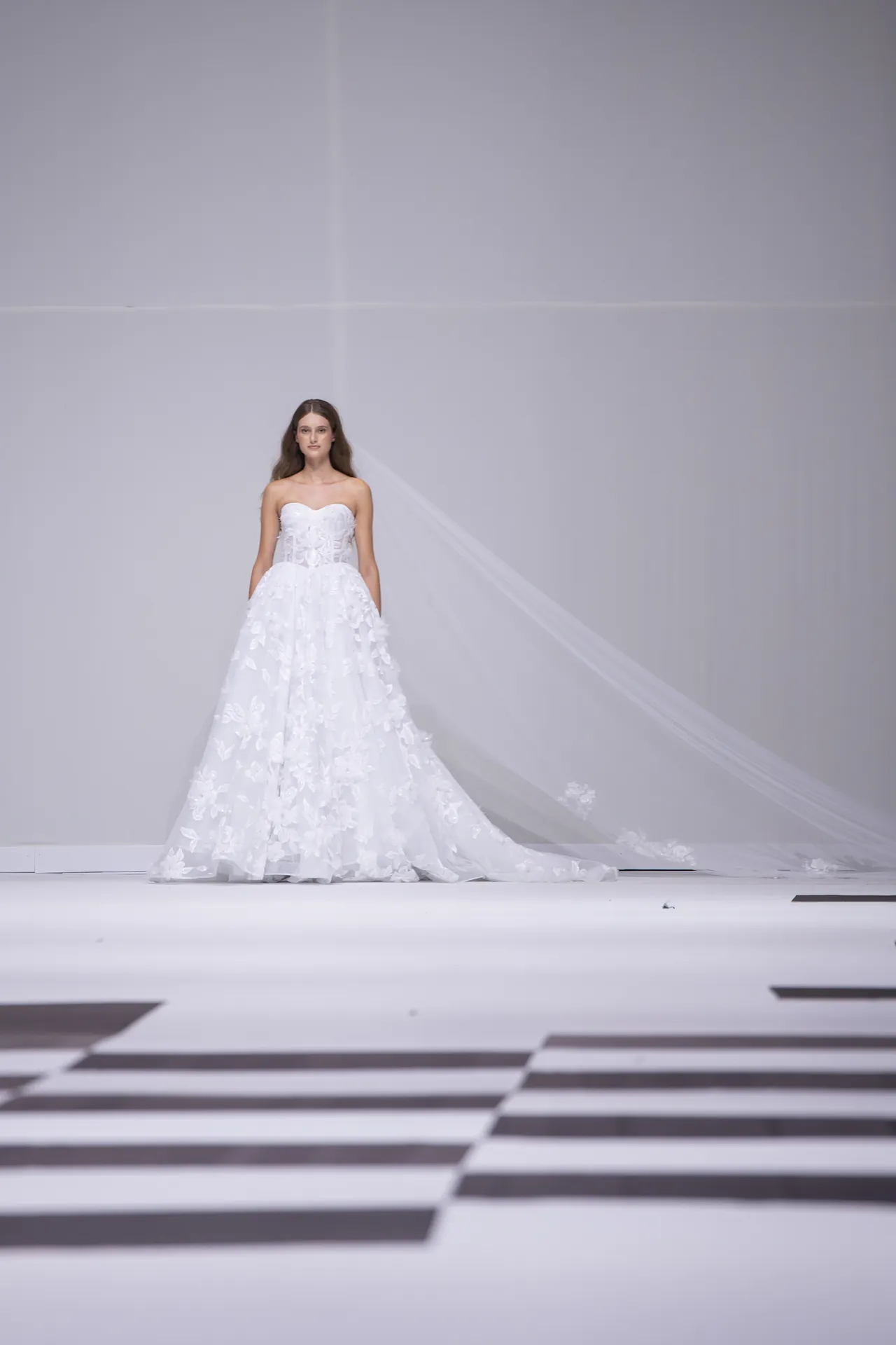 Bride in a strapless white lace wedding gown with a long train and veil walking down a minimalist runway.