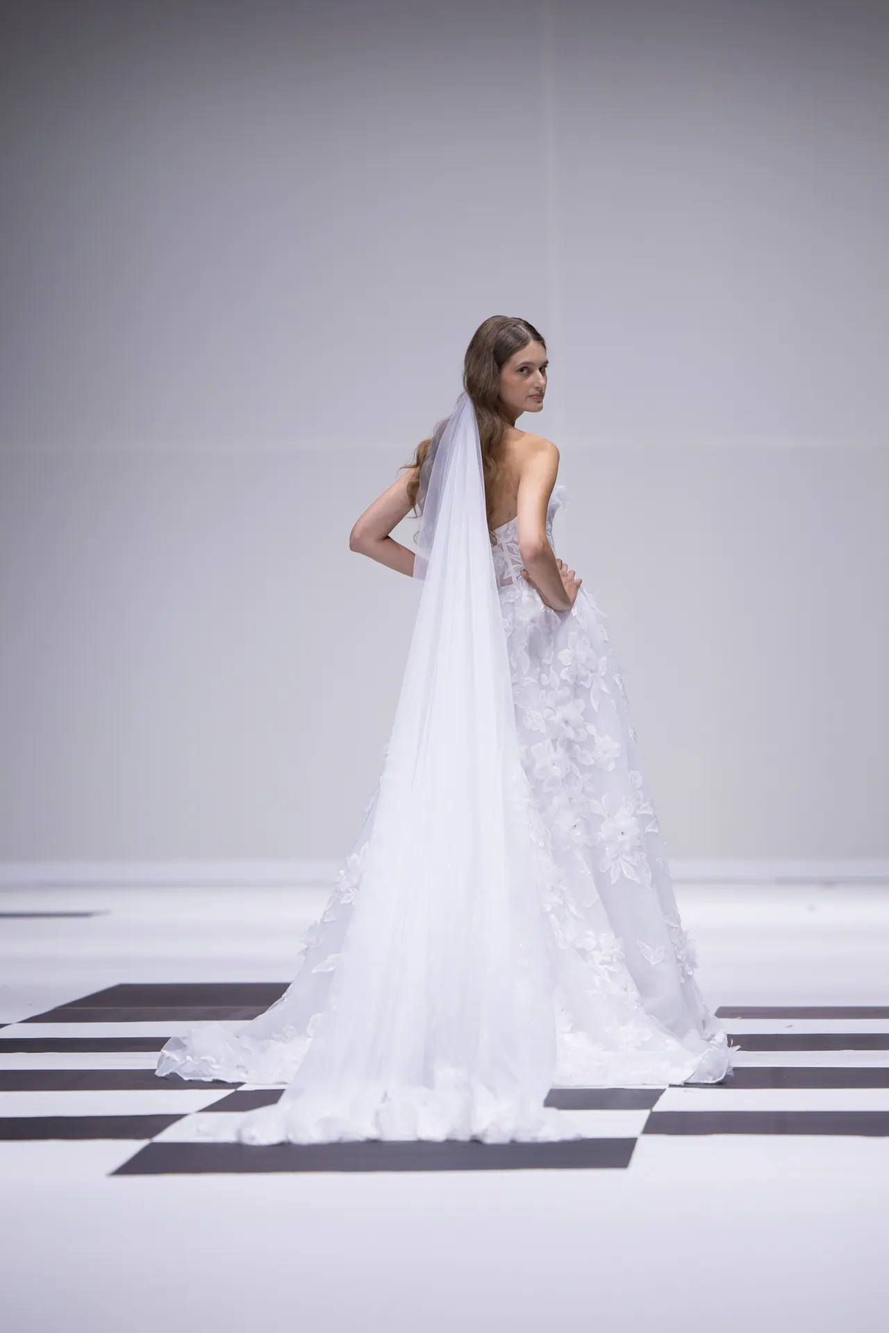 Model in a white wedding gown with a long veil, walking a fashion runway with a black-and-white checkered floor.
