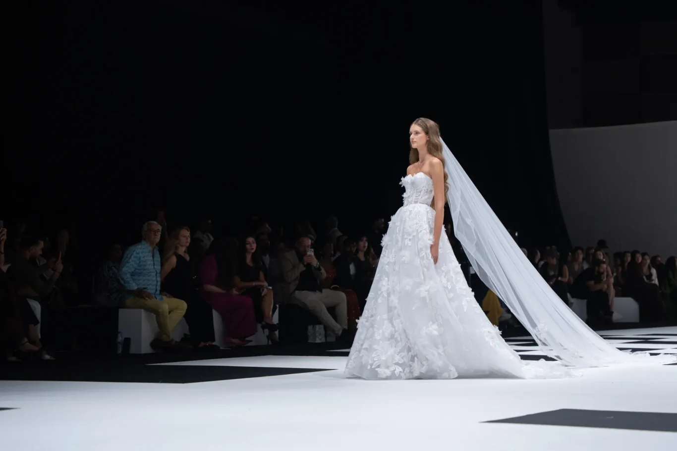 Model in a white bridal gown with long veil walking a fashion-show runway, audience in the background