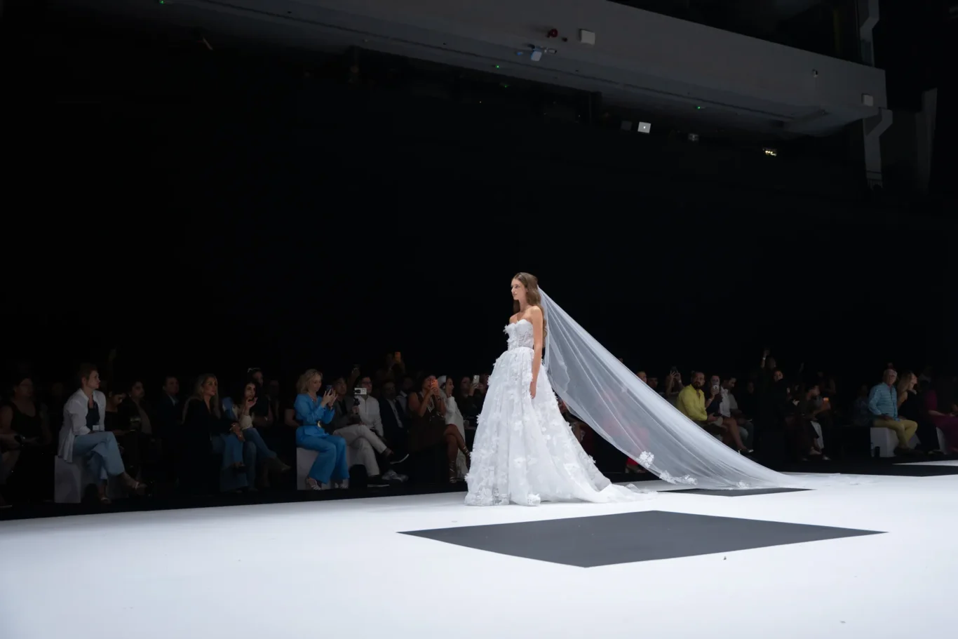 Model in a white wedding gown with a long, flowing veil walks the runway as a seated audience watches the show.