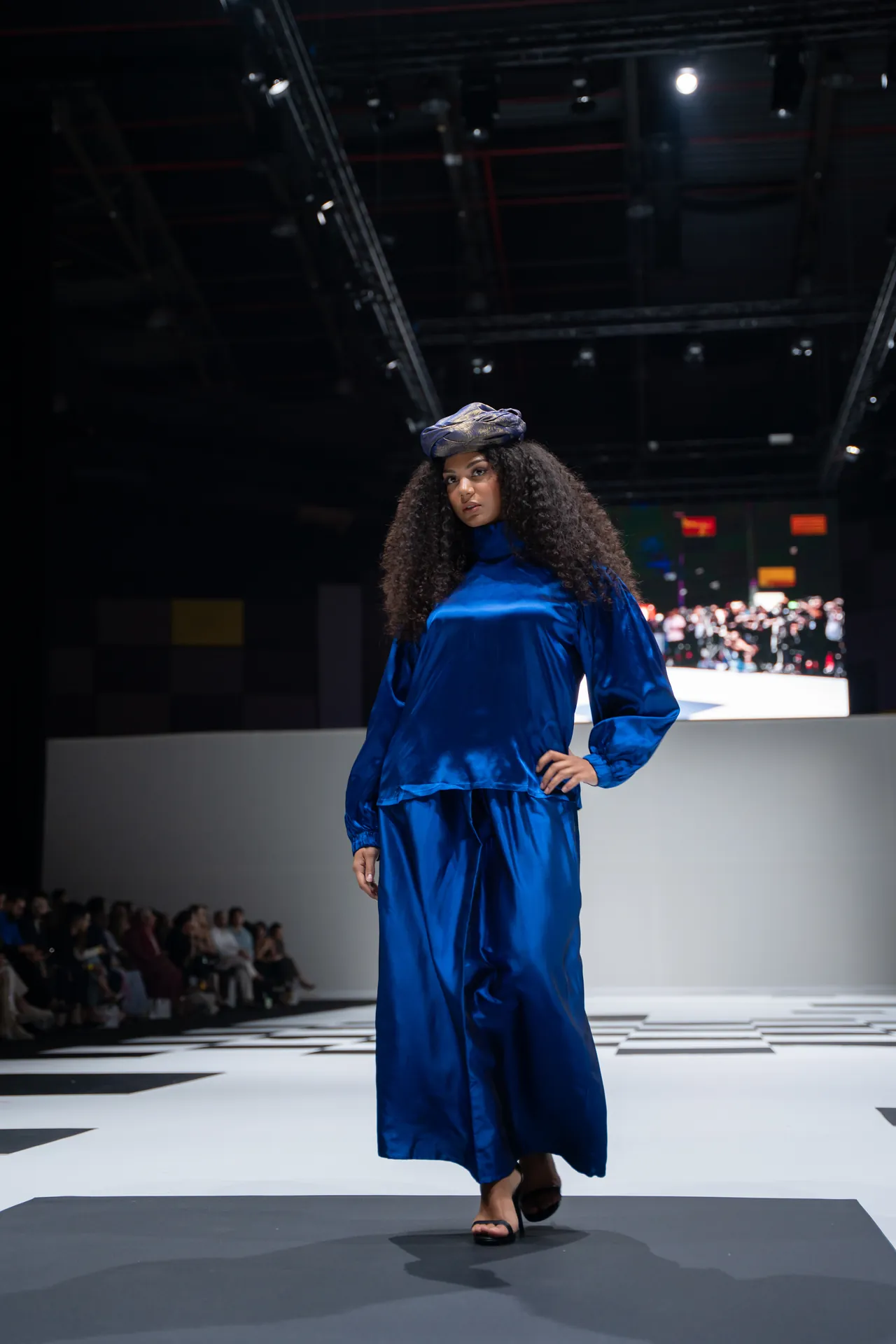Model walks a fashion runway in a cobalt blue satin outfit with a turban-style head wrap; audience sits along the catwalk in the background.