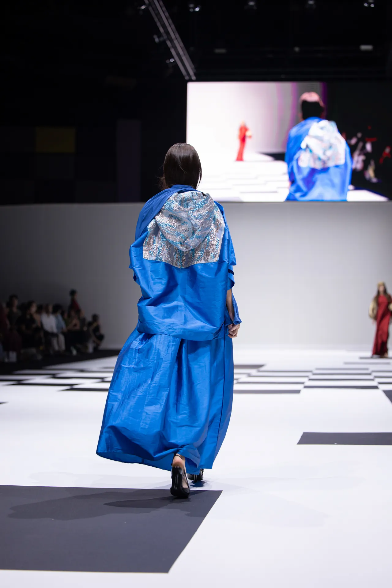 Model in a bright blue, oversized dress walks away on a white runway with black geometric patterns; audience on the left and a large screen shows another model in the background.
