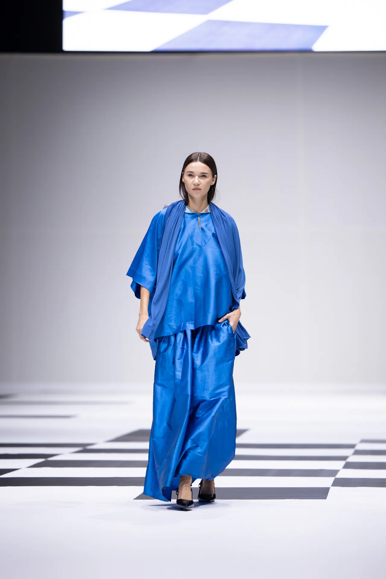 Model walking a runway in a cobalt blue outfit with cape on a black-and-white checkered floor.