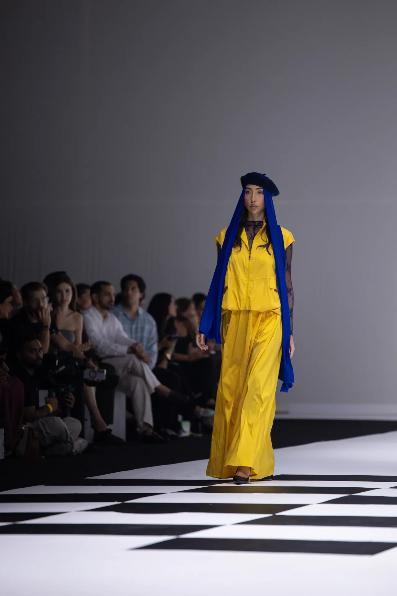 Model walks the runway in a bright yellow outfit with a long blue scarf and navy beret, audience visible in the background.