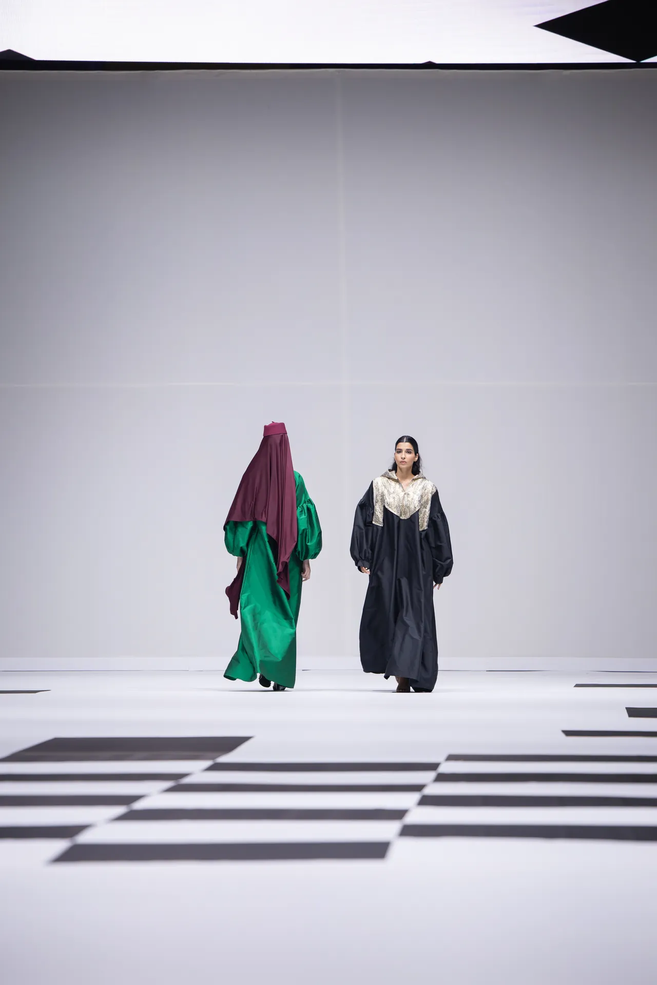 Two fashion models walk a minimalist runway in oversized gowns, one green and purple and the other black with metallic detailing, against a stark geometric floor.
