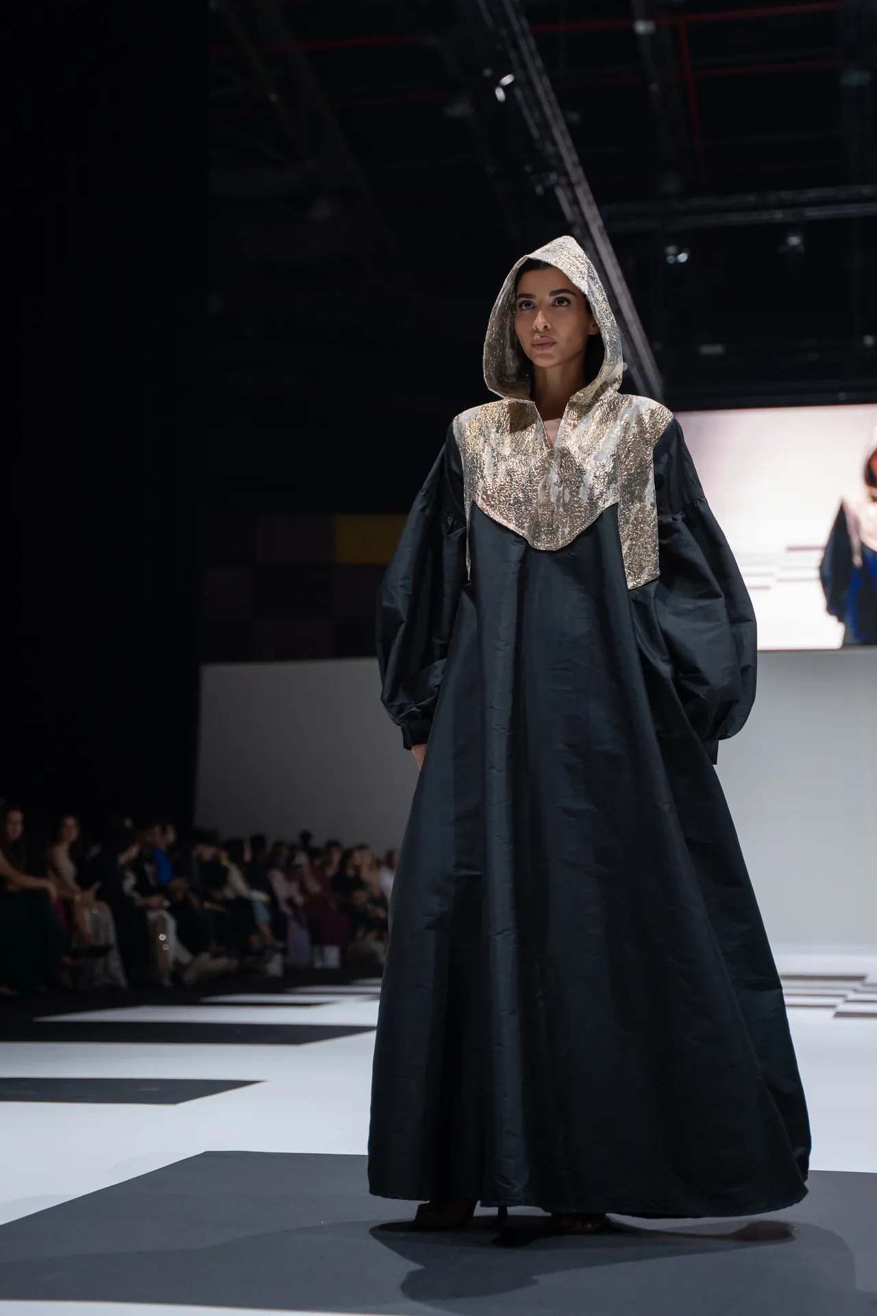 Model walks a fashion show wearing a long dark gown with a gold sequined hood and bib detail, on a striped runway.