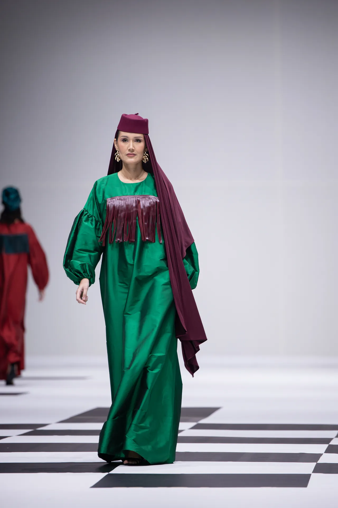 Model walking runway in a green dress with a burgundy headwrap and long scarf, on a black-and-white checkered catwalk.