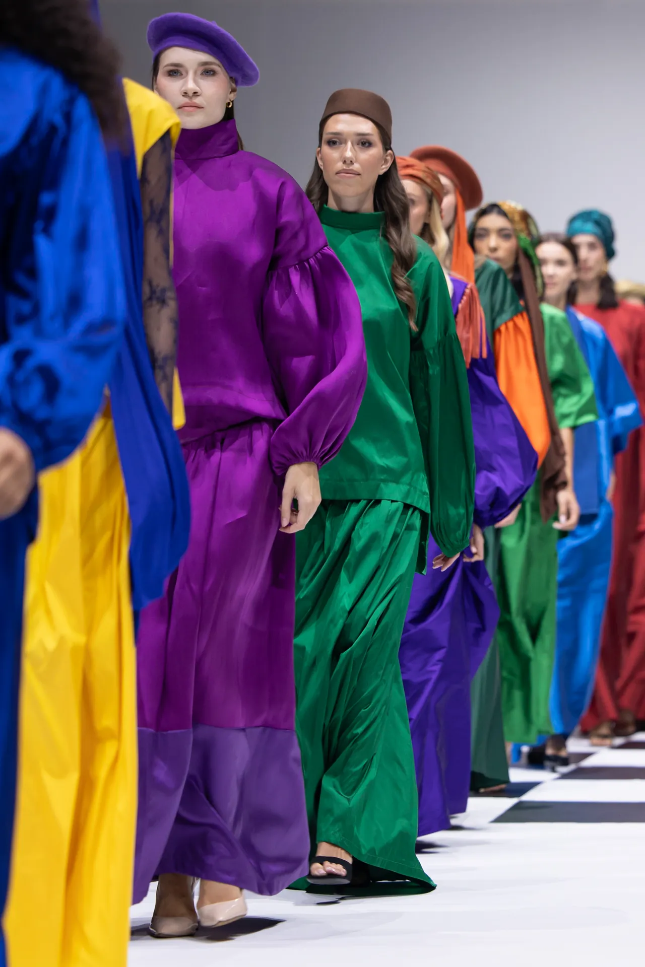 Models in bright, flowing gowns and matching hats walk a runway in a rainbow lineup at a fashion show