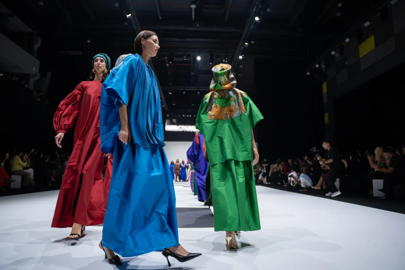 Models walk a fashion runway wearing oversized, colorful gowns (red, blue, green) with an audience watching from the sides under bright stage lights.
