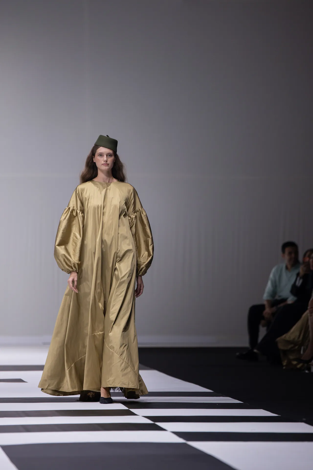 Model walks a runway in a long metallic gold gown with oversized puff sleeves and a green military-style hat, on a black-and-white striped catwalk.