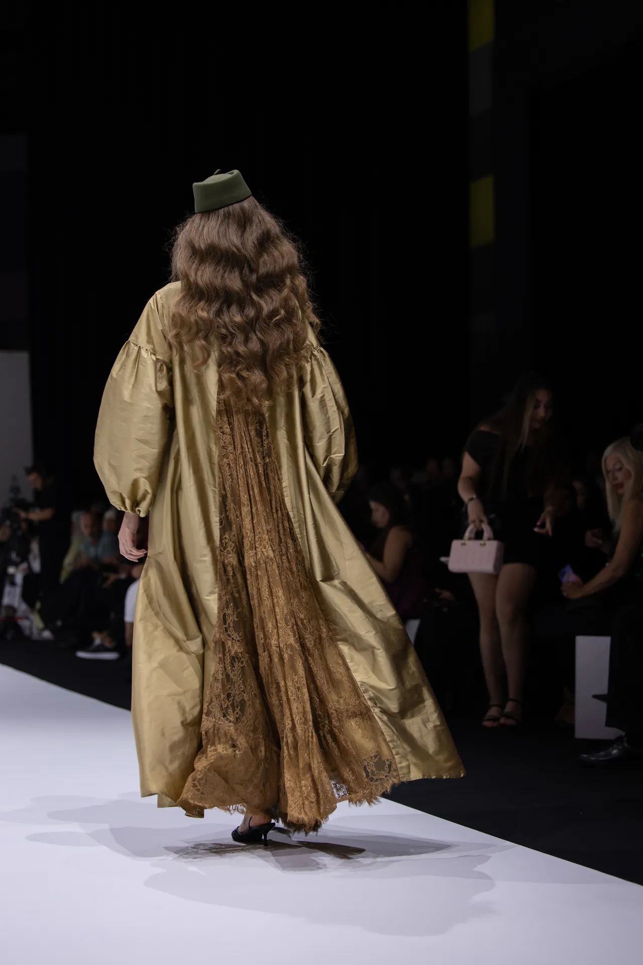 Back view of a model walking the runway in a gold gown with lace panels; long wavy hair and a green hat; audience in the background.
