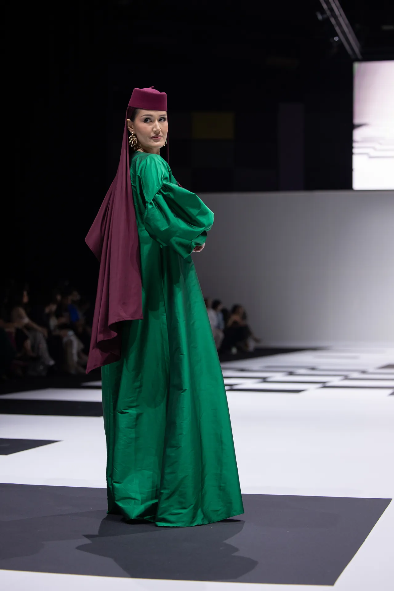 Model walking a fashion show in a bright green gown with a magenta headpiece and cape on a runway.