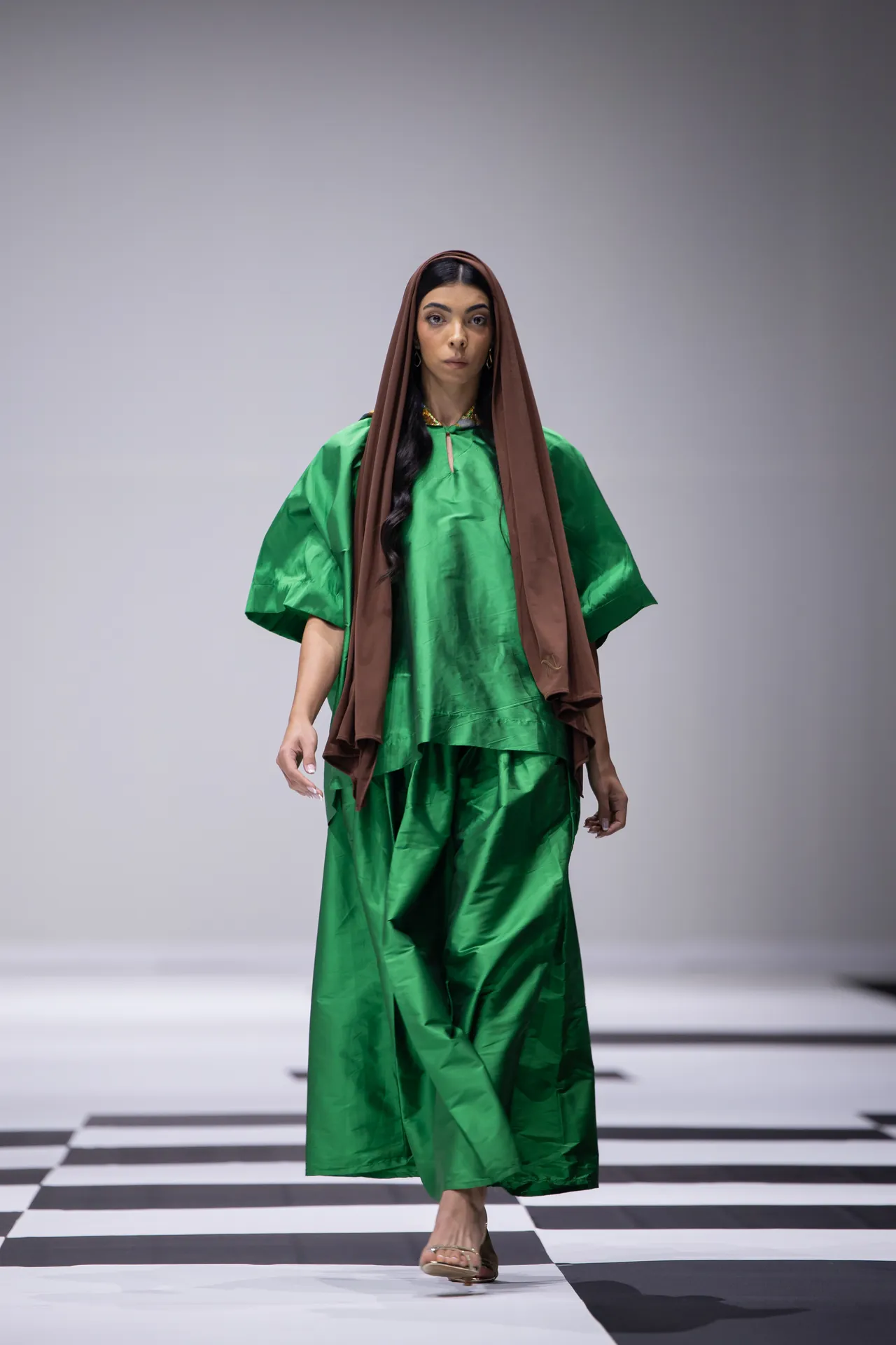 Model walking a fashion runway in a bright green flowing outfit with a brown headscarf against a neutral backdrop, on a black-and-white checkered floor.