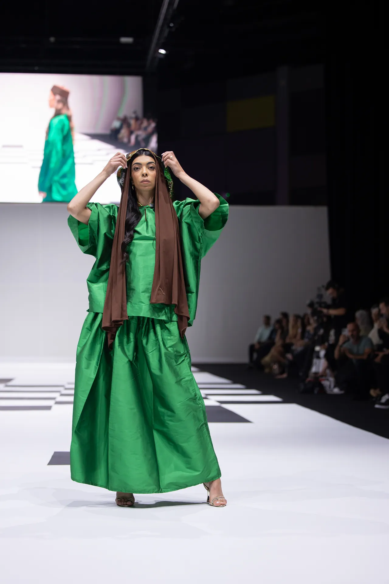 Model walks a white runway in a vivid green gown with a brown scarf, facing the camera; audience watches in the background.