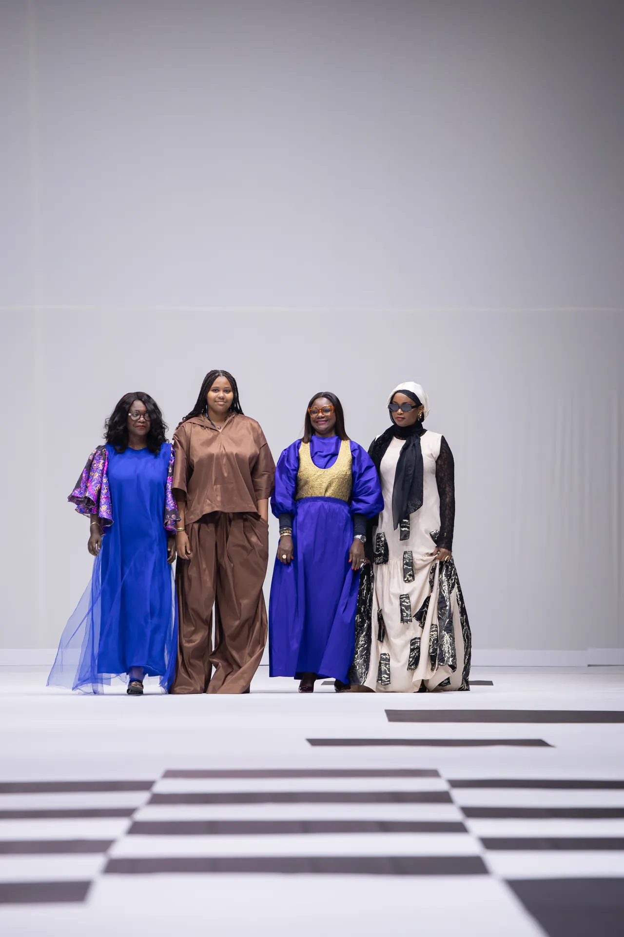Four models walk a runway in vibrant outfits on a black-and-white checkered floor.