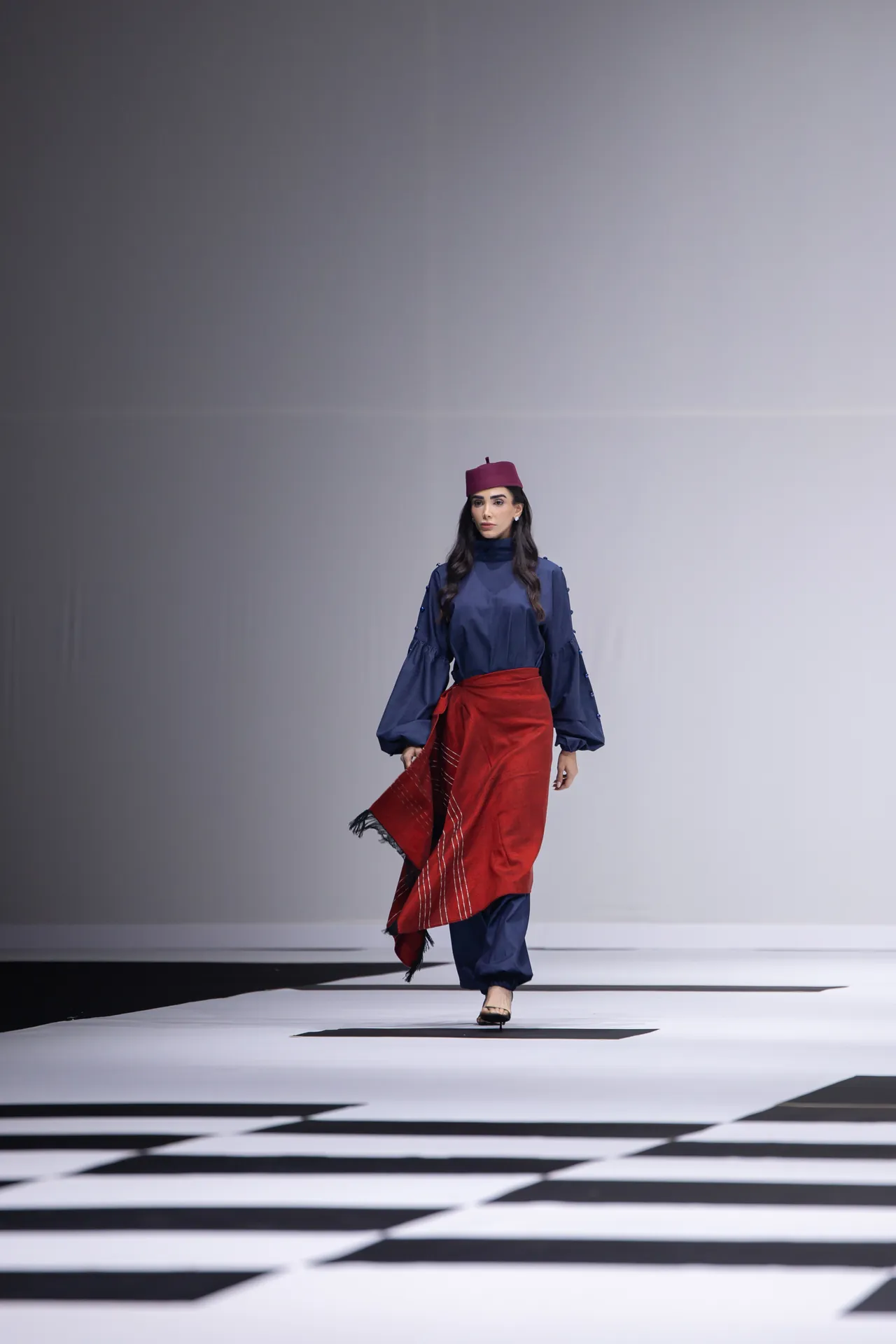 Model walking a fashion runway in a navy blue ensemble with a red sash and magenta hat against a gray backdrop