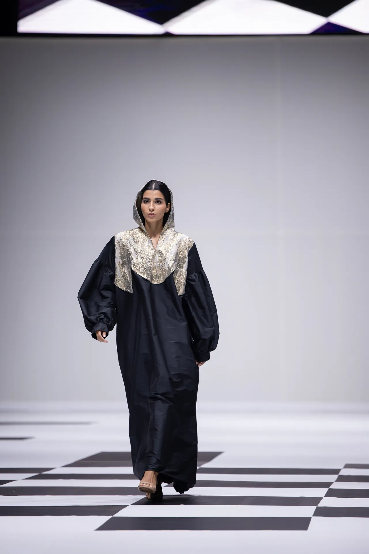 Model walking a fashion runway in a loose black dress with a gold-sequin hood on a black-and-white checkerboard catwalk.