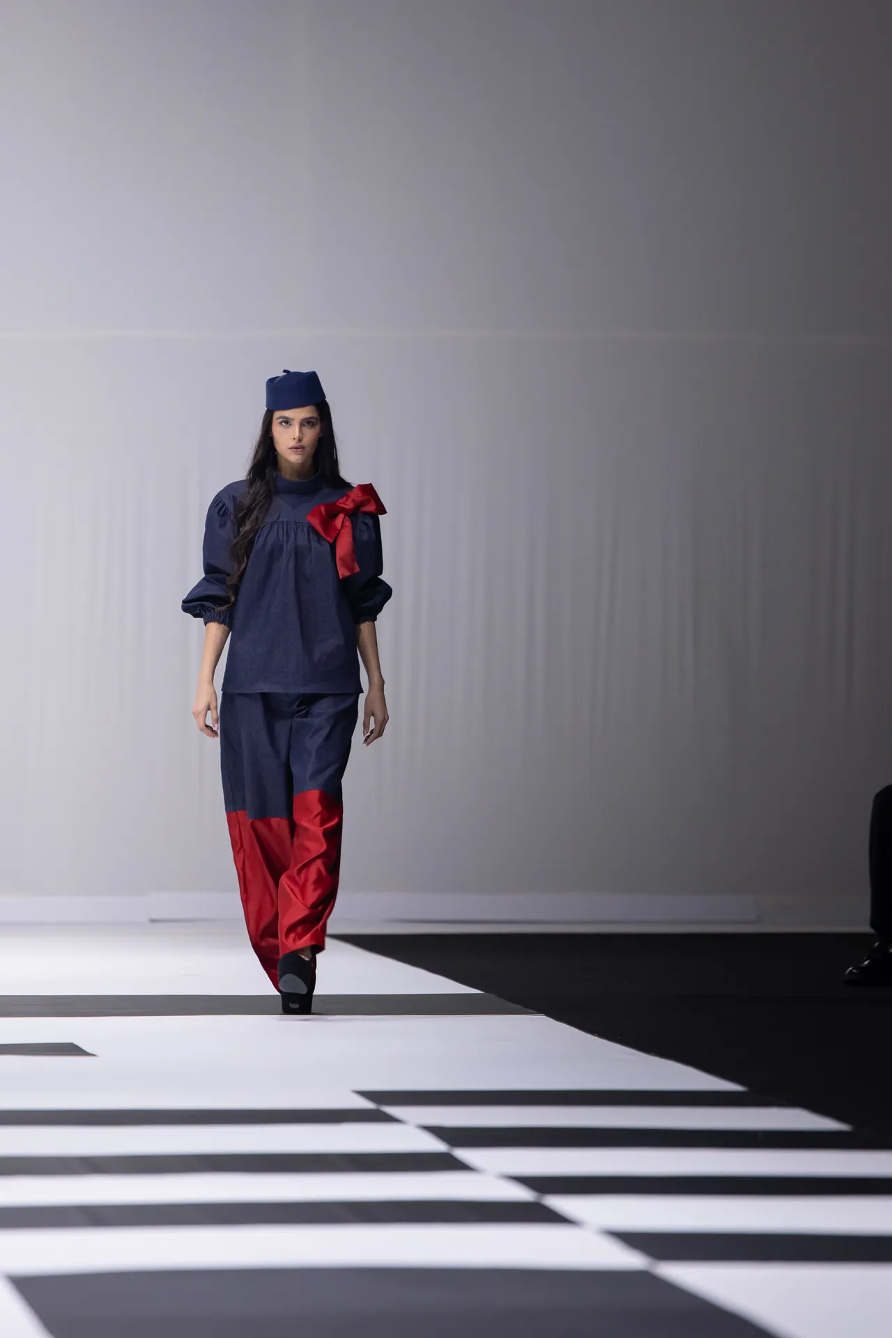 Model walking a fashion runway in a navy outfit with a red bow and matching blue cap on a minimalist stage.