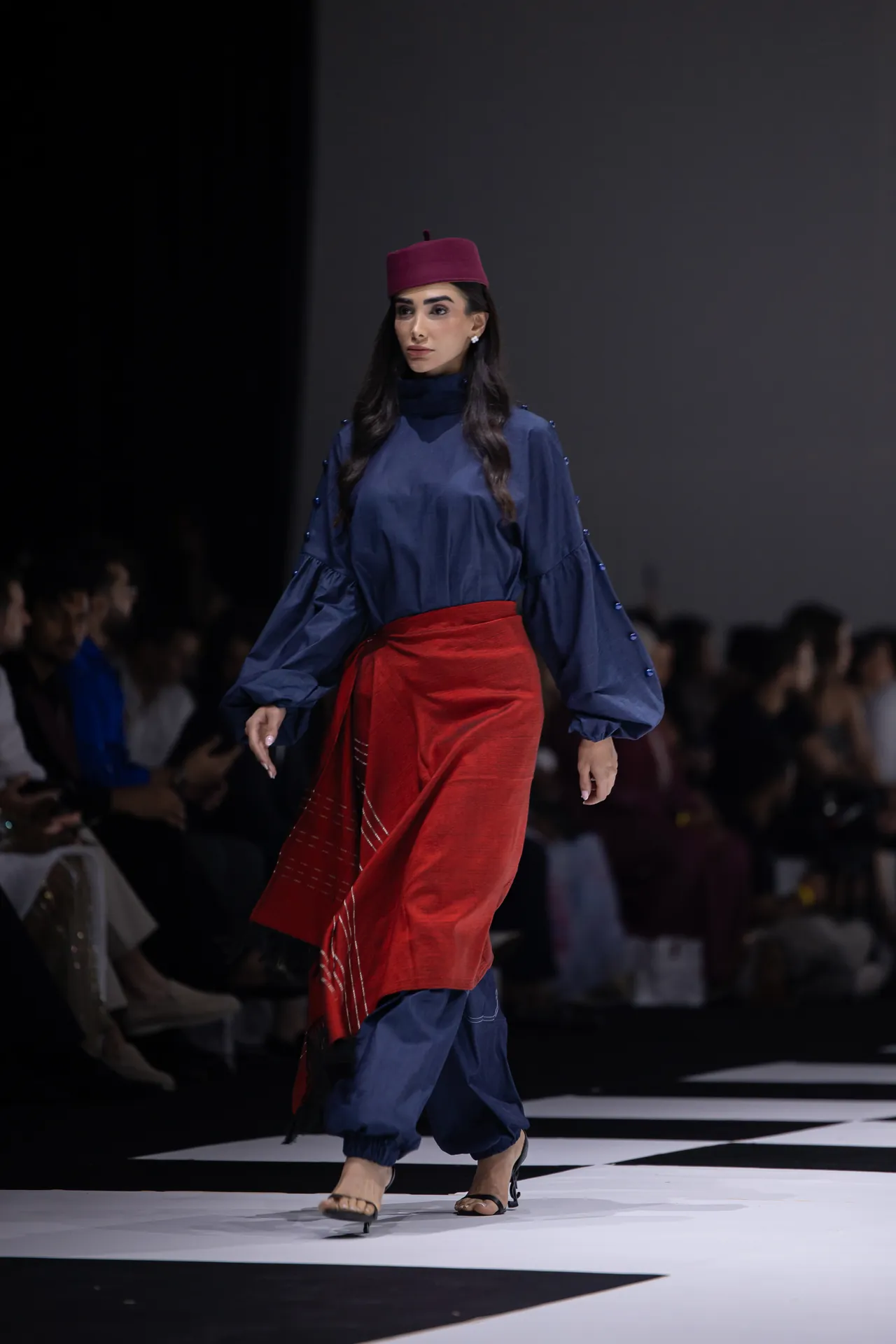 Model walking a fashion show runway in a navy blue outfit with a red wrap skirt and a magenta hat, audience blurred in background.