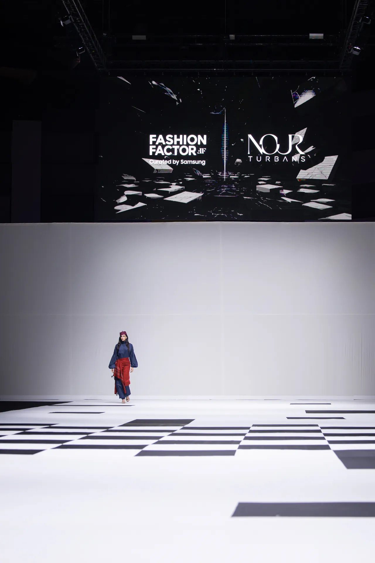 Model walks down a white runway with a black-and-white checkerboard floor and a large screen displaying 'FASHION FACTOR' and 'NOR TURBANS' behind her.