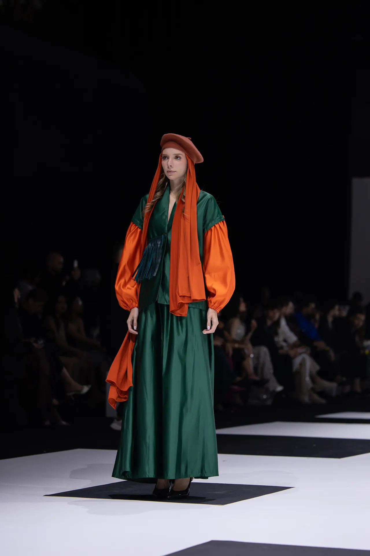 Model walking a fashion runway in a green satin gown with bright orange sleeves and a matching orange beret, audience in the background.