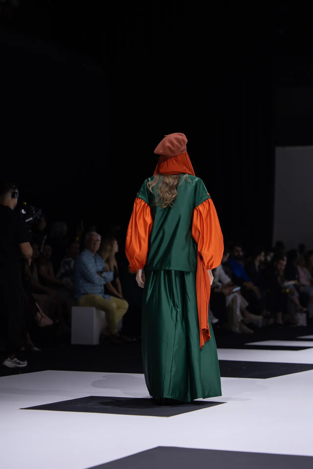 Model walking away on a fashion runway in a green gown with bright orange puff sleeves and an orange headscarf, seen from behind.