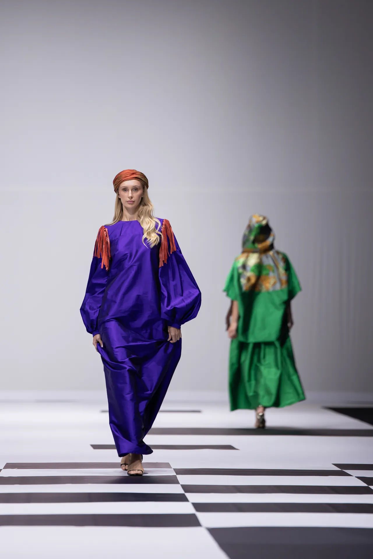 Model walks the runway in a bold purple oversized outfit with an orange headscarf; another model in green is visible in the background on a checkered floor.