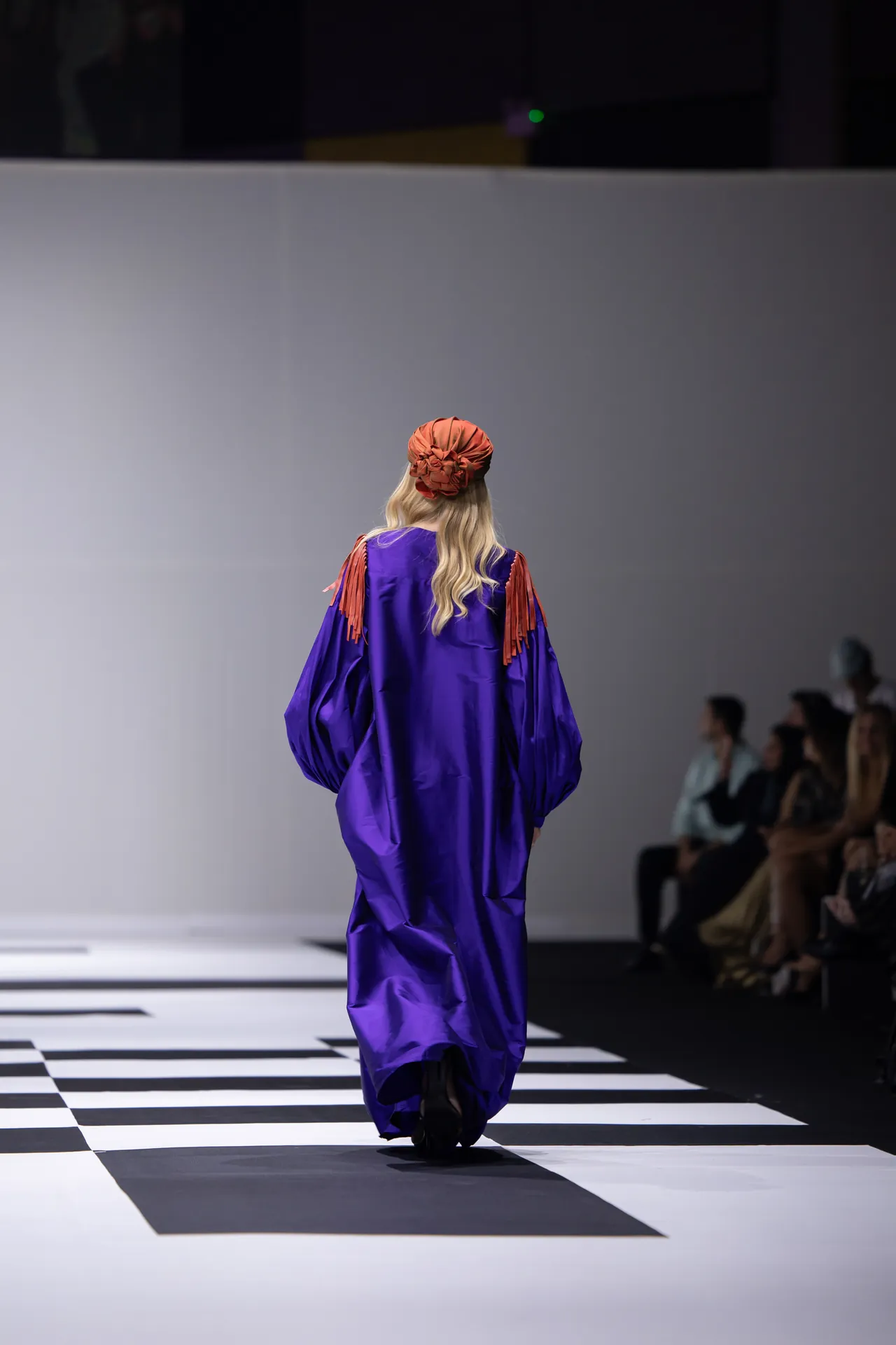 Model walks away on a checkered runway wearing a loose purple gown with orange tassel shoulders and an orange scrunched headpiece, audience visible to the right.