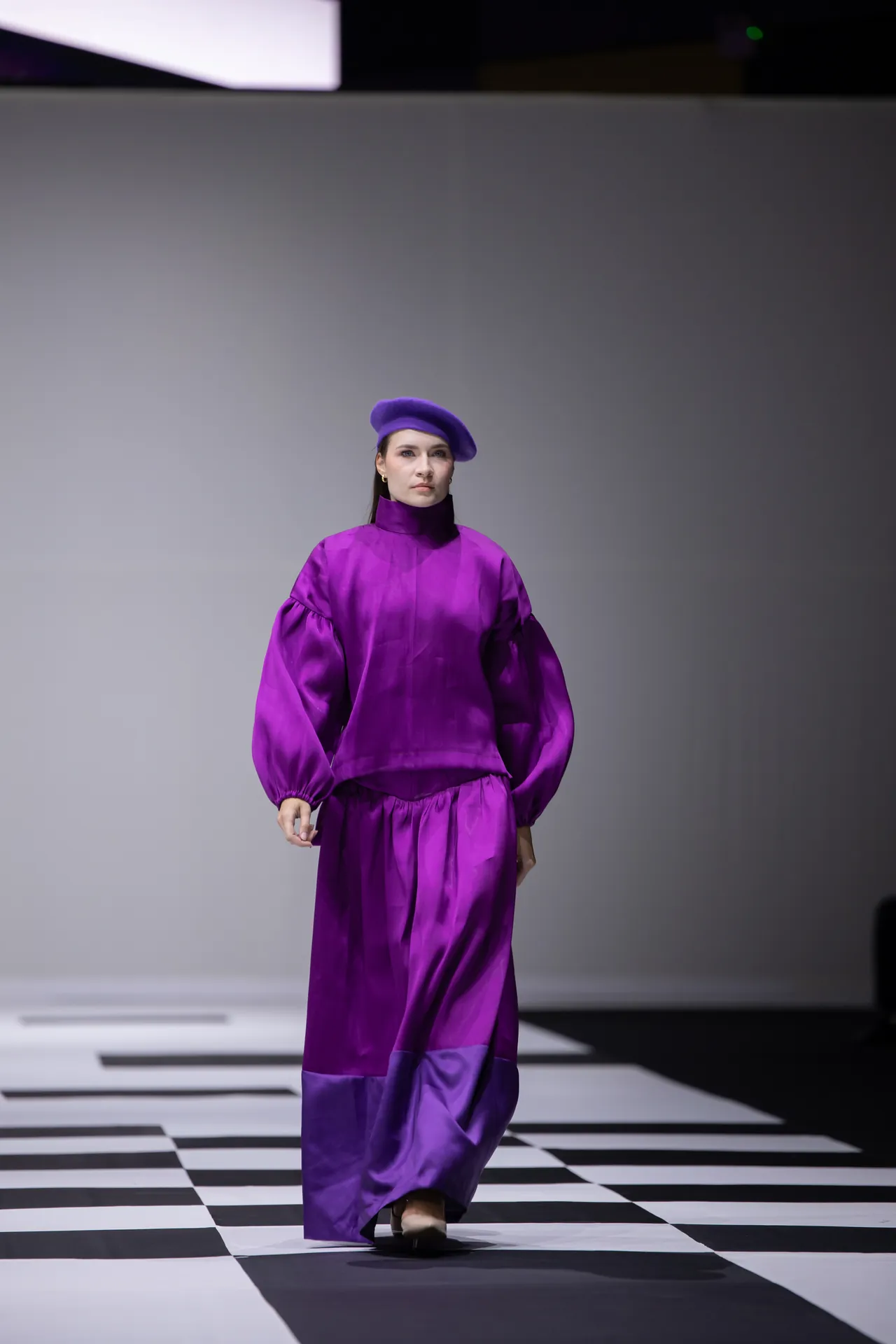 Model wearing a vibrant purple outfit and beret walking down a black-and-white checkered runway.